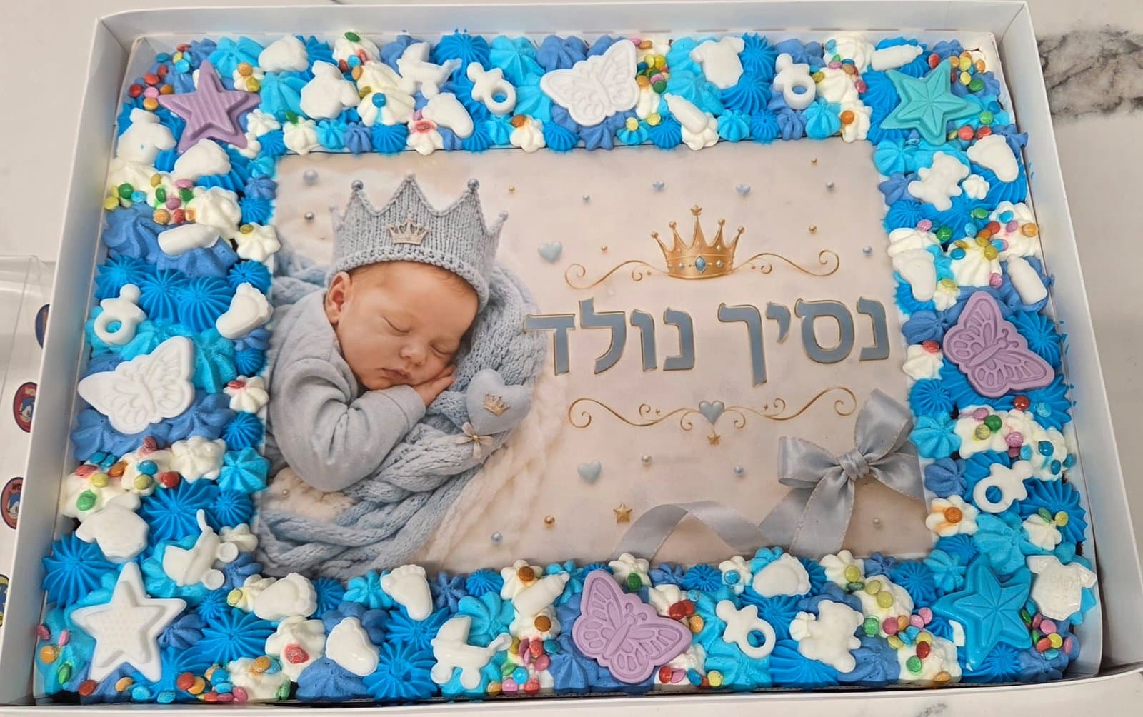 דף סוכר עם תינוק ישן בכתר סרוג וכיתוב נסיך נולד, מסגרת קצפת כחולה ופרפרים